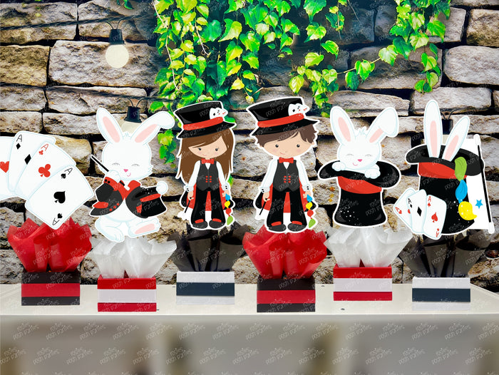 Kids birthday party magician themed centerpieces. The centerpieces feature illustrated cutouts including playing cards, a white rabbit in a tuxedo, a girl magician, a boy magician, a rabbit popping out of a top hat, and a magician's hat with a wand. Each cutout is set in a red, white, or black base filled with matching tissue paper. Receive fully assembled like pictured.
