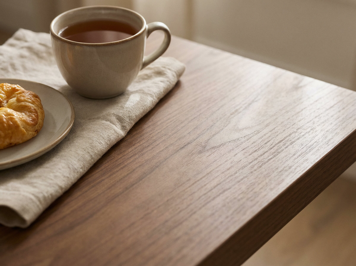 A warm, close-up lifestyle shot of a wooden table with a light beige ceramic cup of tea on a matching napkin, alongside a small plate with a croissant.