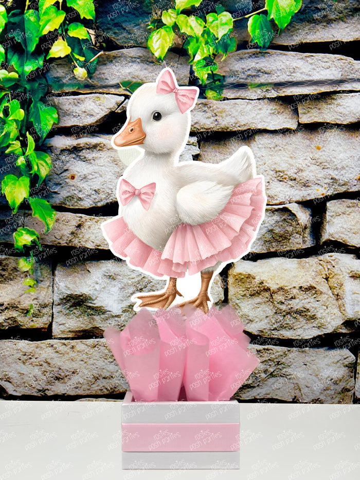 fully assembled party centerpieces featuring adorable watercolor-style white baby geese or ducklings dressed in pink outfits. The designs include a goose in pink boots, a pink sunhat, a pink cowboy hat, a pink tutu, a large pink bow, and one inside a pink floral frame. Each cutout stands on a white base wrapped in pink ribbon with fluffy pink tissue paper.